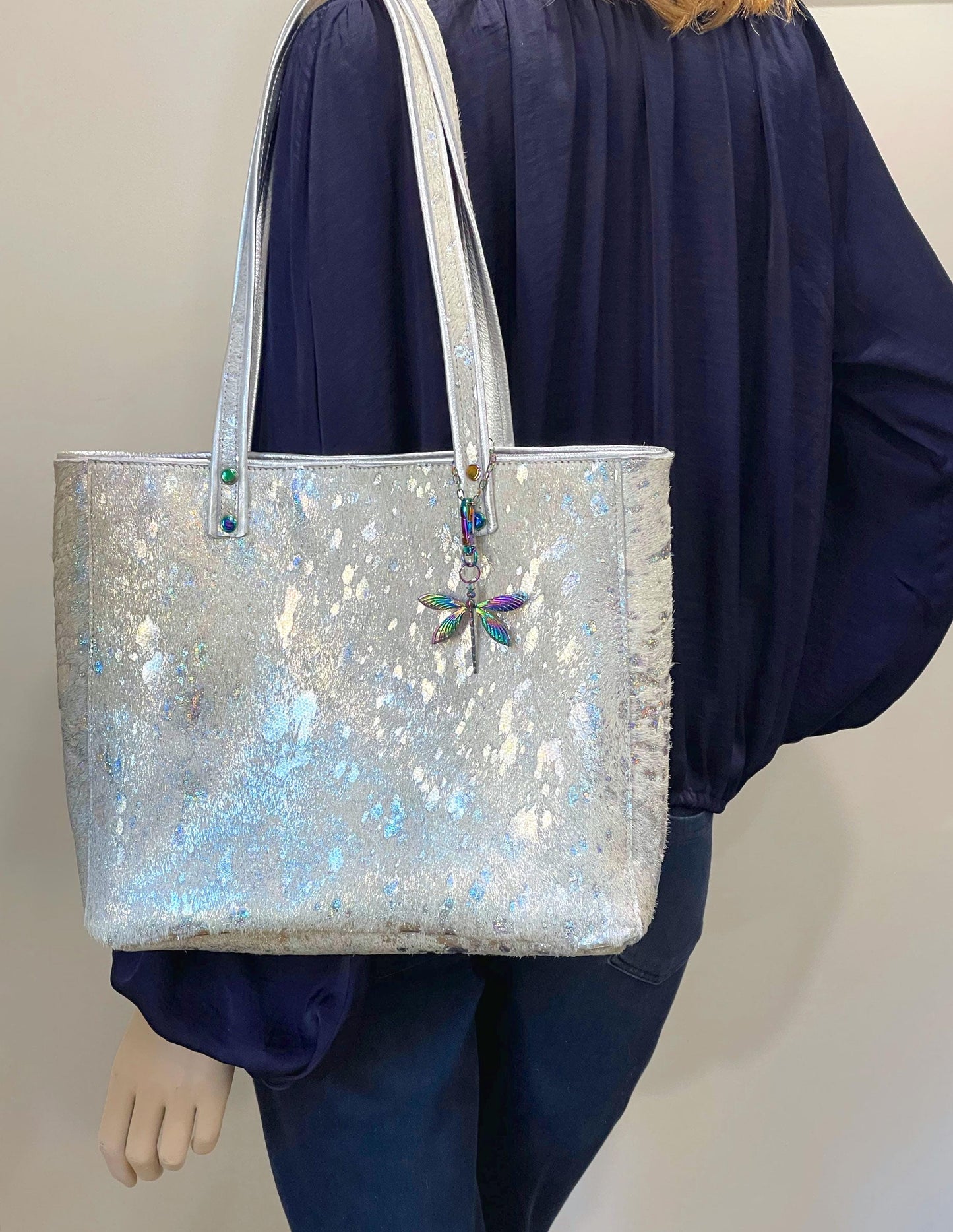 Iridescent silver acid wash cowhide tote bag with a dragonfly charm held by a person wearing a blue outfit.