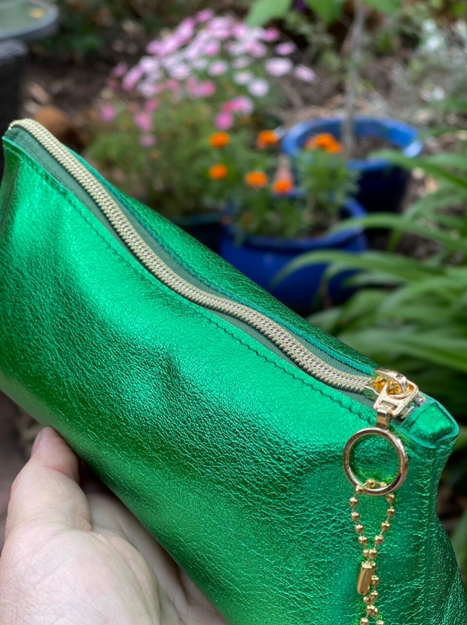 Metallic green leather zipper pouch with gold zipper and ring pull held in front of potted plants.