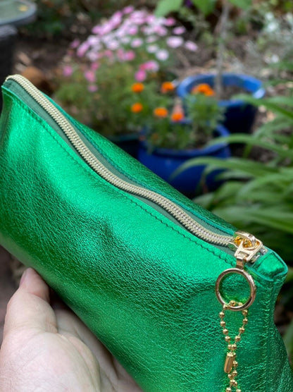Metallic green leather zipper pouch with gold zipper and ring pull held in front of potted plants.