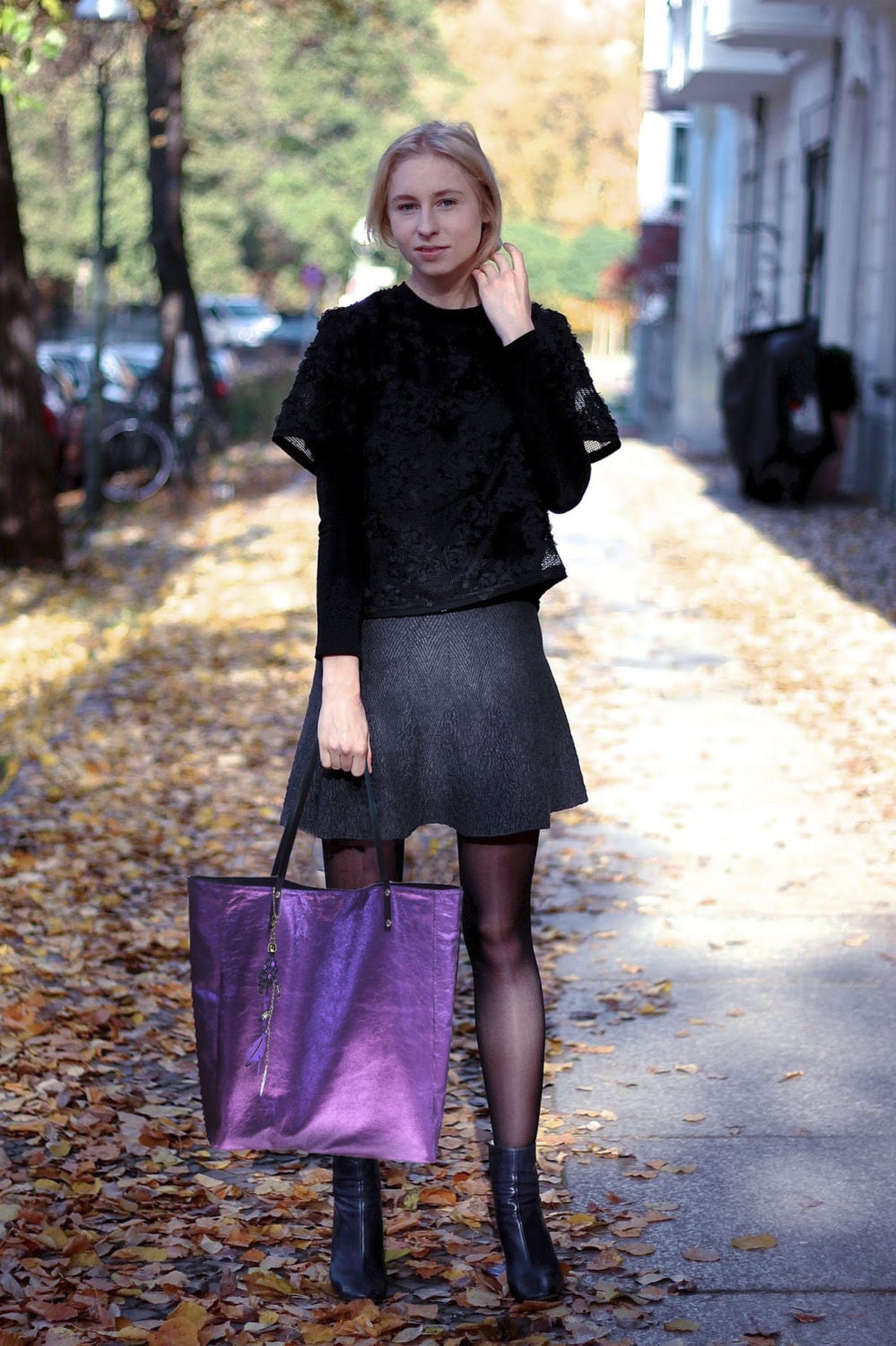 Woman in black outfit with a purple bag walking on a leaf-covered street.