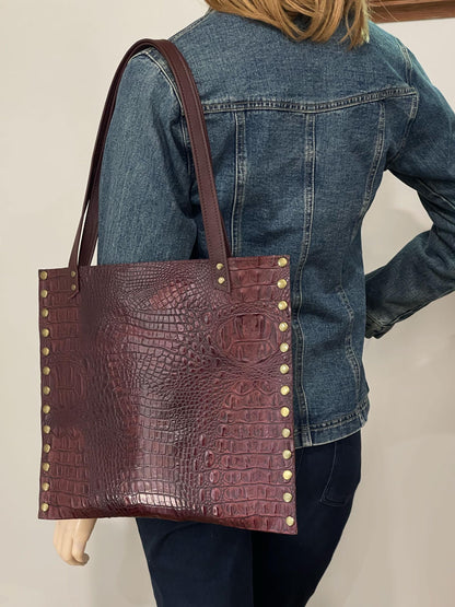 Person wearing a denim jacket with a Burgundy tooled leather tote bag featuring crocodile texture and gold studs.