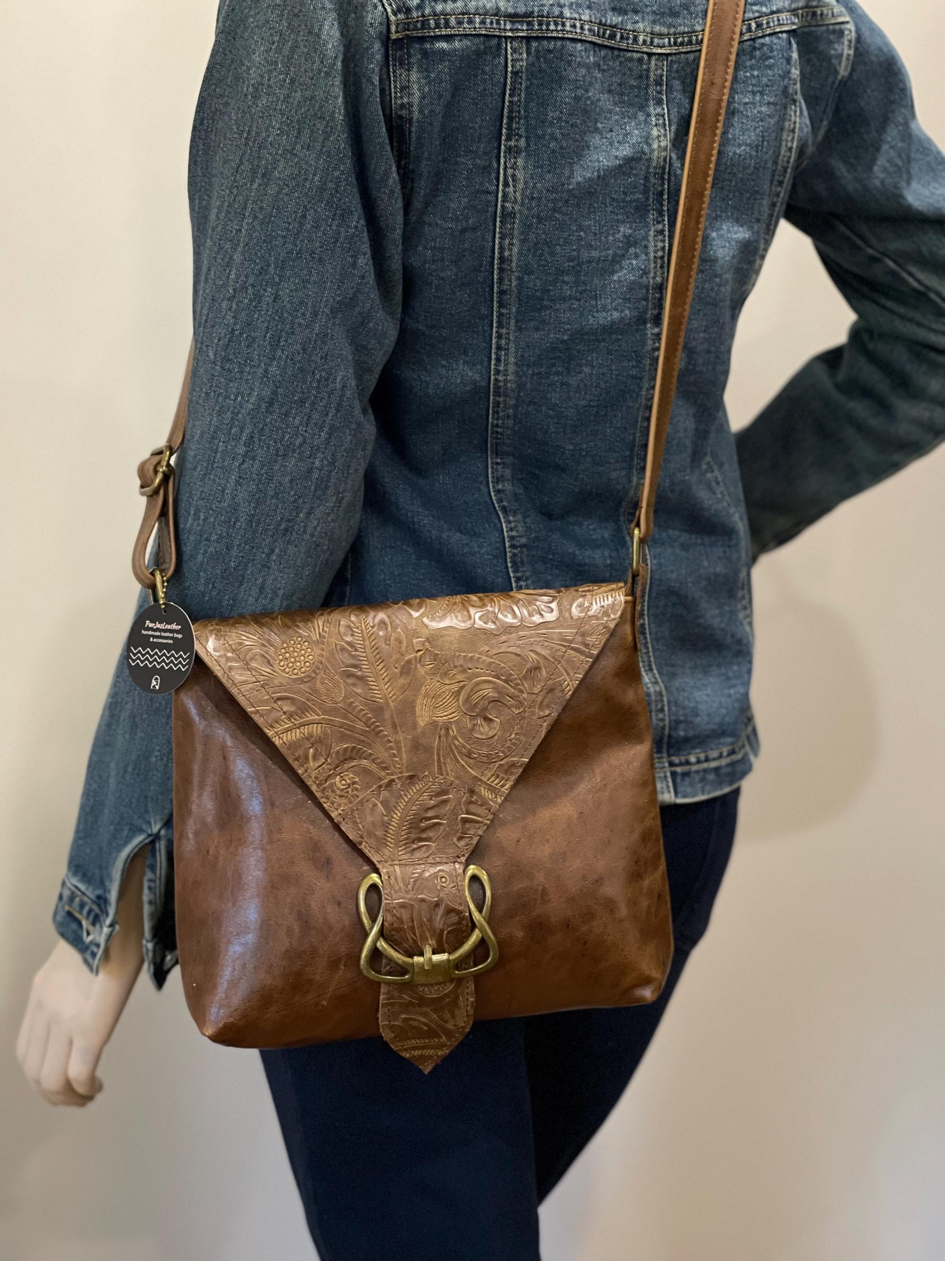 Person wearing a denim jacket with a brown tooled leather bag with celtic style buckle over their shoulder against a neutral background
