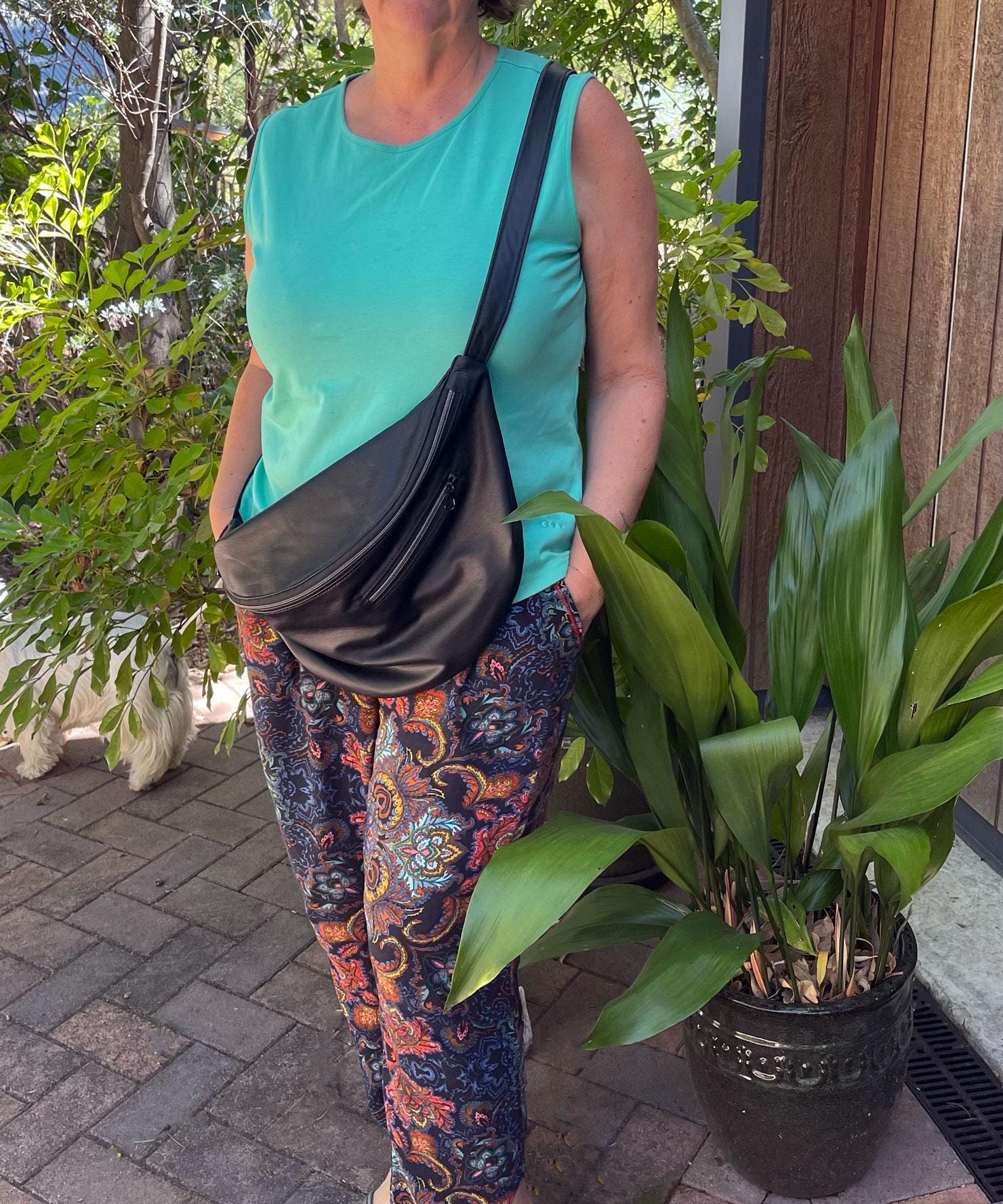 Person with crossbody black half moon sling bag that has outside pocket, standing next to a potted plant outdoors.