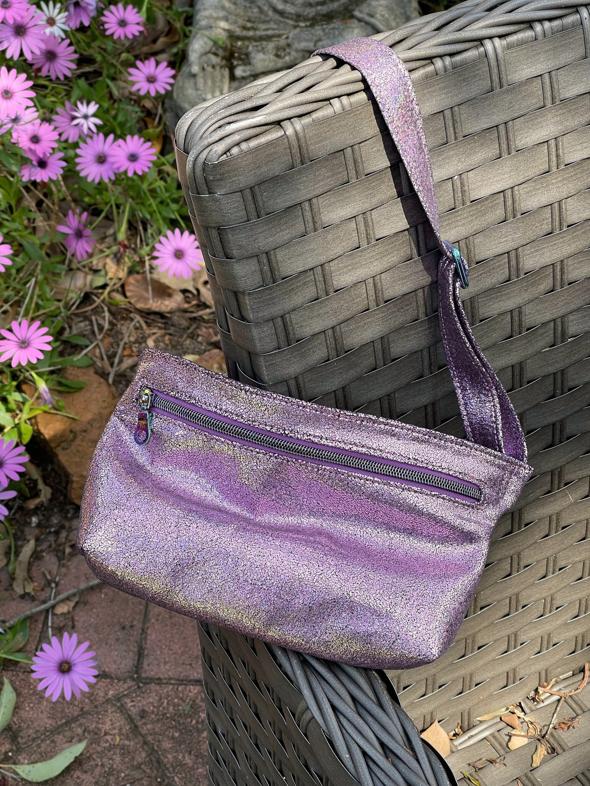 Pink glittery bum bag with purple zipper and rainbow hardware with purple flowers in the background