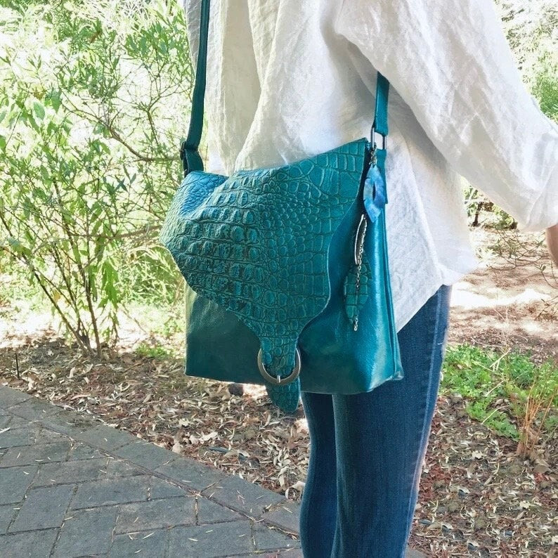 Person wearing a teal handbag with a textured croco patterned flap against white blouse and jeans.