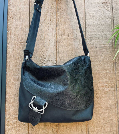 Black leather handbag with embossed sloped flap closure and celtic style buckle featured on a wooden surface