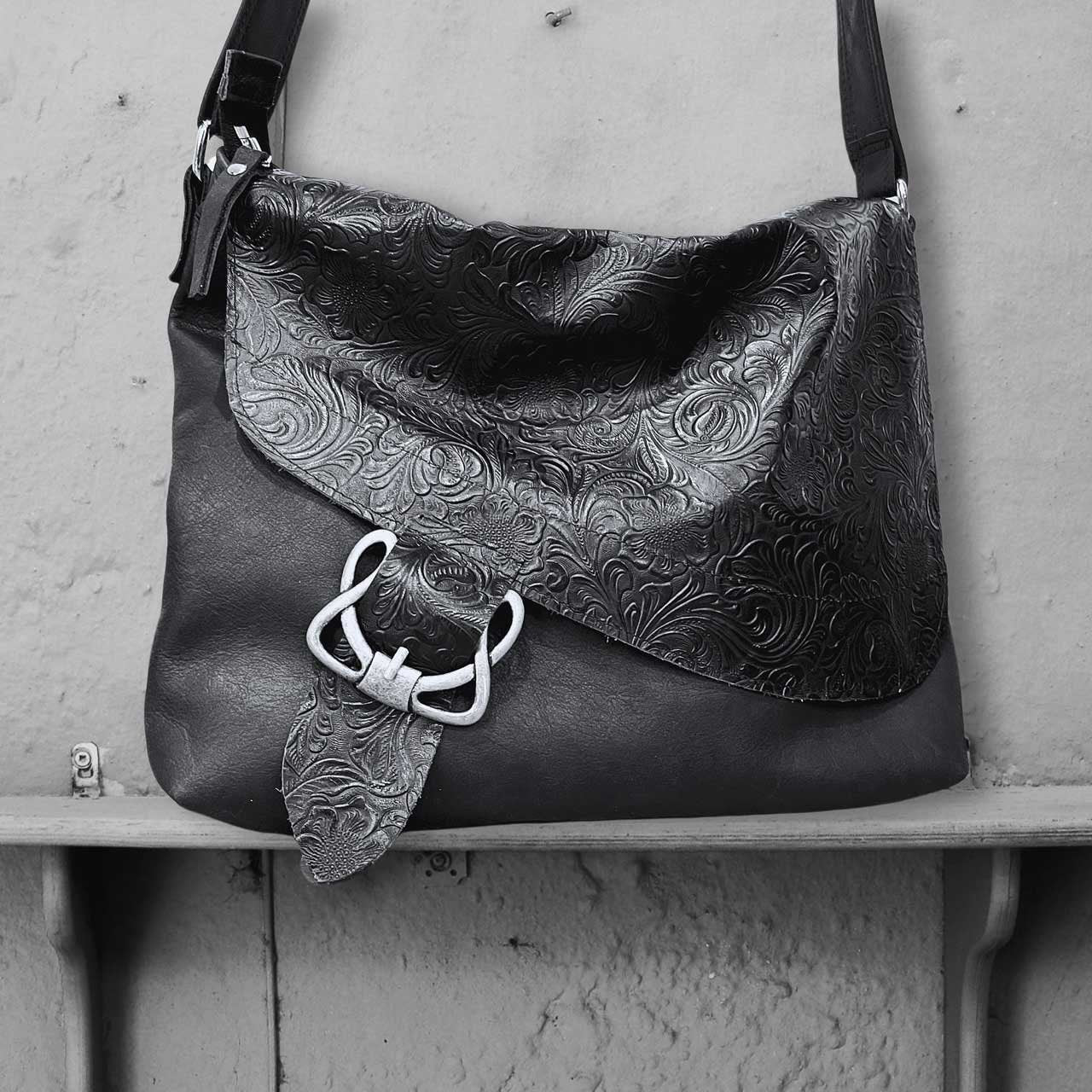 Black leather handbag with intricate patterns and a decorative celtic metal buckle clasp on a gray surface.