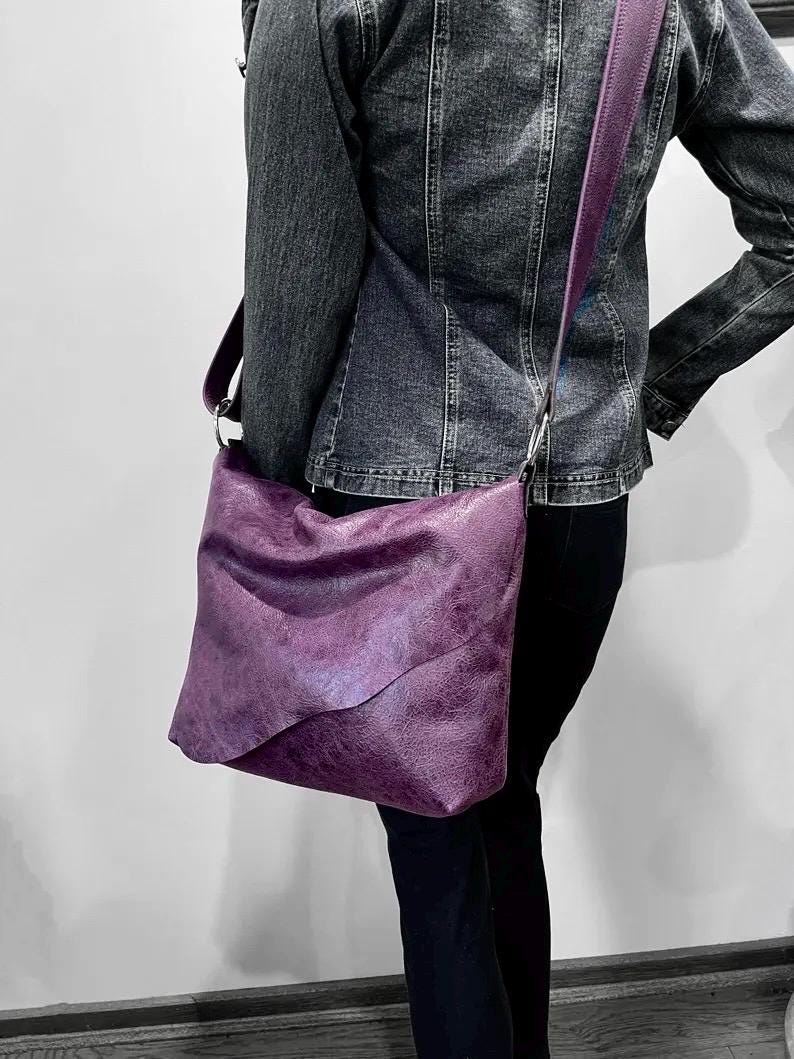 Person wearing a denim jacket with a distressed vintage purple shoulder bag with irregular long flap on a plain background