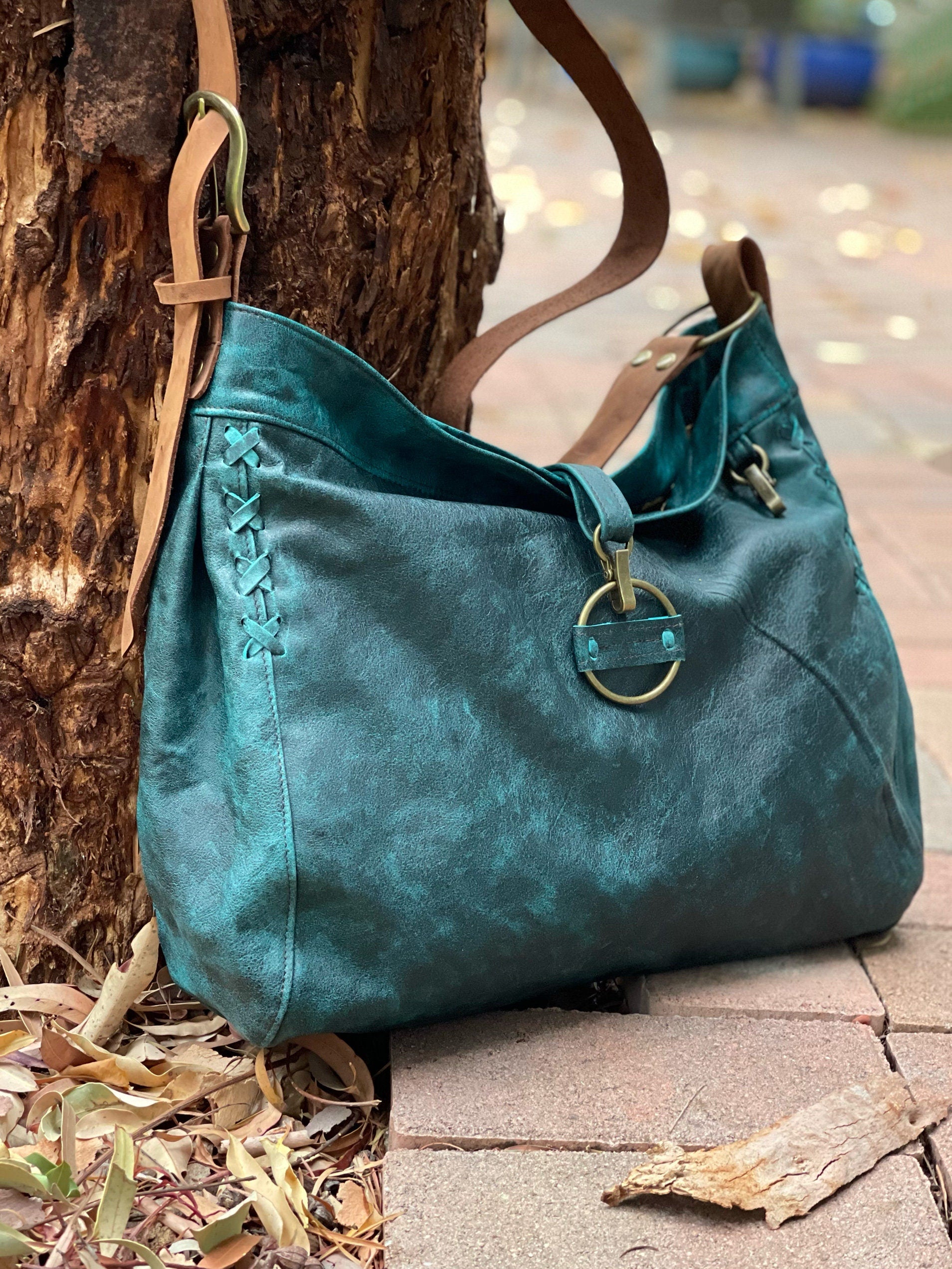 Teal handbag with brown leather strap and lacing and pleat leaning against a tree on a stone path.