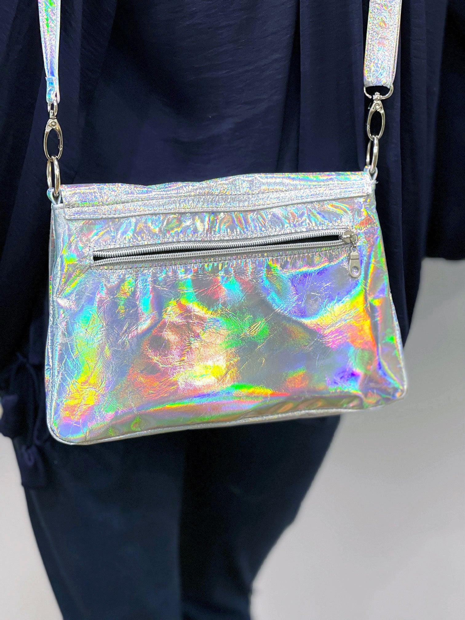 Holographic handbag with reflective aurora colour surface and zipper pocket held by a person wearing dark jeans.