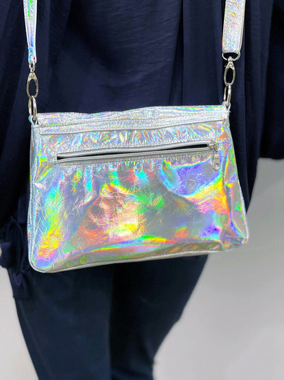Holographic handbag with reflective aurora colour surface and zipper pocket held by a person wearing dark jeans.
