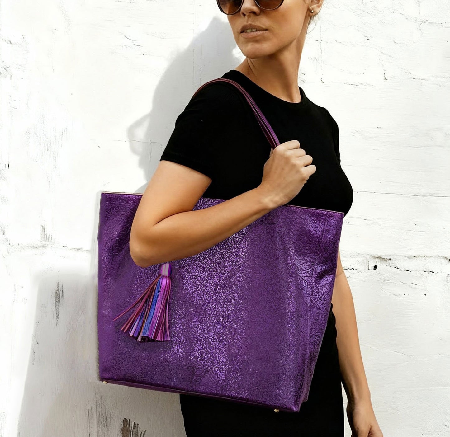 Person holding a purple tote bag with tassels against a white brick wall. 2 shoulder straps. protective feet on bottom