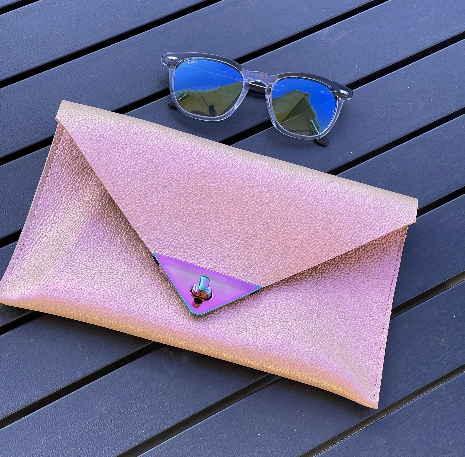 Pink clutch bag with a metallic clasp on a wooden surface, accompanied by a pair of sunglasses.