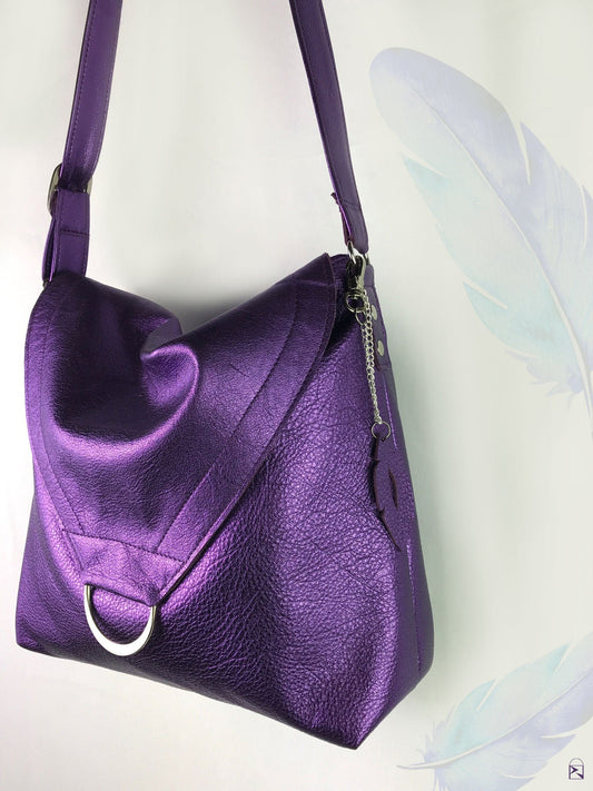 side view of metallic Purple leather handbag with flap and ring feature, and a cut leather charm on a white background