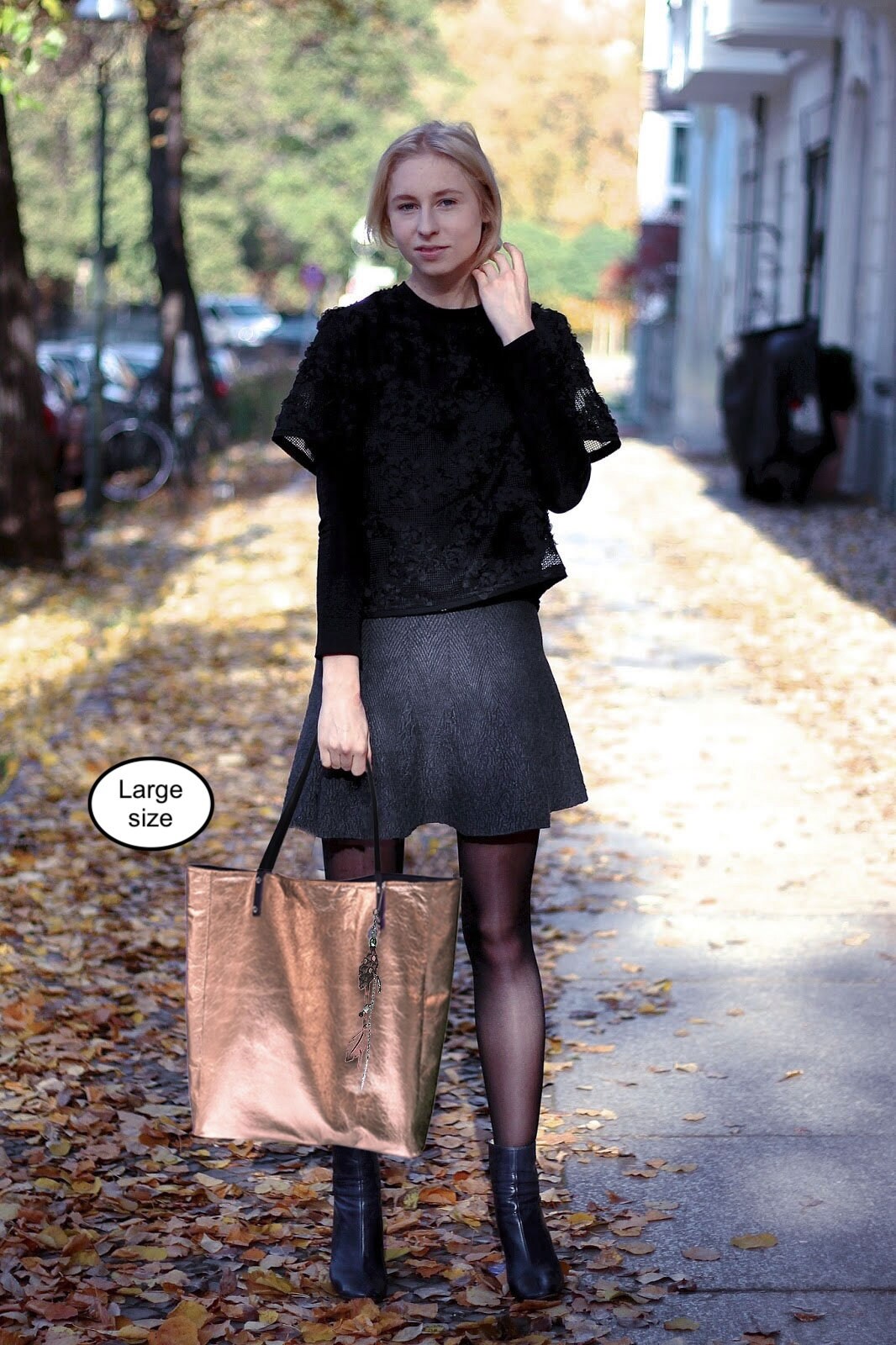 Woman in black outfit holding a rose gold leather tote  bag on a leafy street.