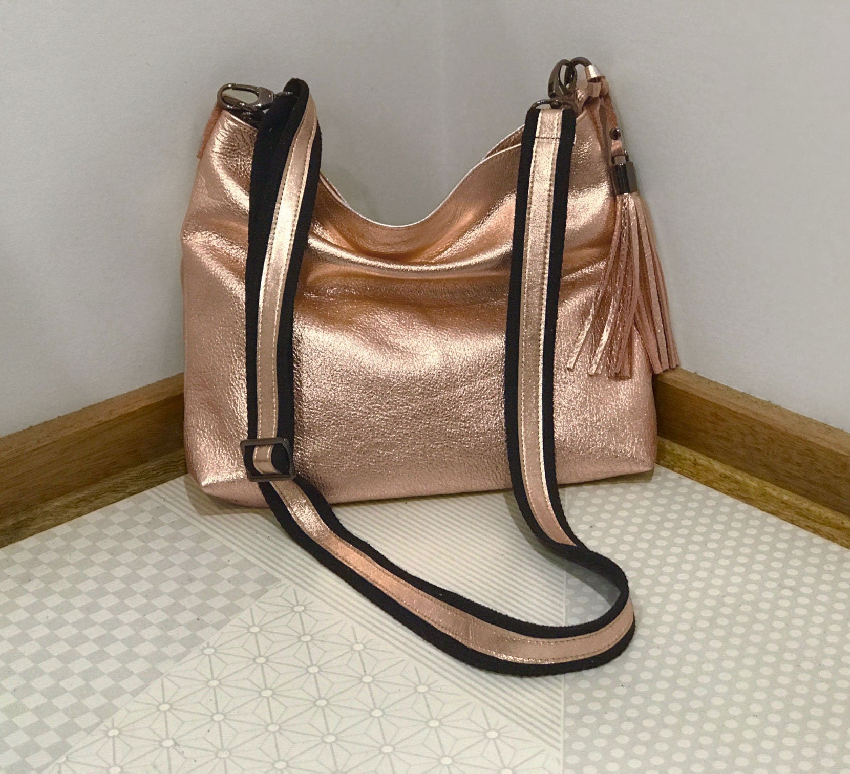 Rose gold handbag with a black and rose gold strap and matching tassel on a light gray floor.