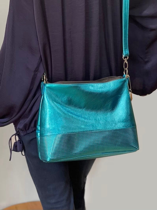 Teal metallic handbag with textured detail worn over crossbody as hands free bag against a neutral background