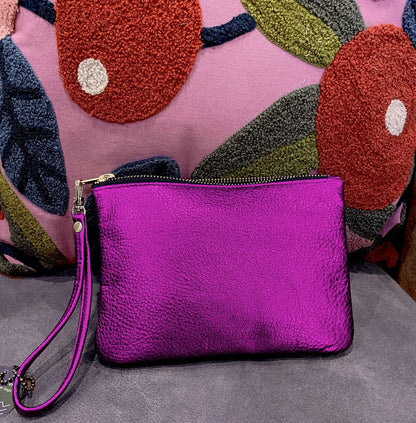 fuchsia pink metallic leather clutch bag with a wrist strap on a gray surface with a colorful pillow in the background.